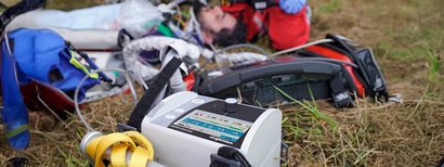 A WEINMANN ventilator lies on a meadow in the foreground. Rescue workers attend to an injured person.