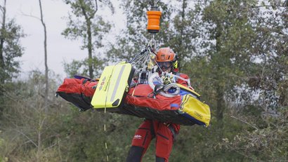 Ein Luftretter wird mit einem Patienten in einer Trage per Seilwinde aus einem bewaldeten Gebiet geborgen.