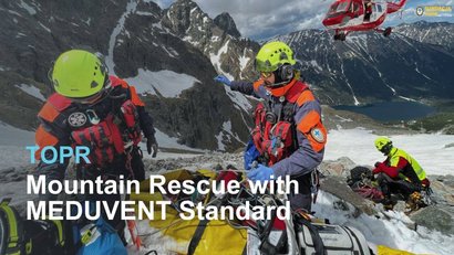 Mountain rescuers with helmets and equipment help an injured person in the snow. A helicopter flies in the background. A ventilator lies on the ground.