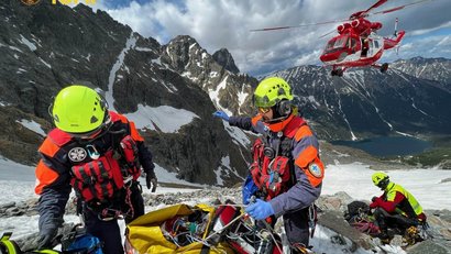 Mountain rescuers with helmets and equipment help an injured person in the snow. A helicopter flies in the background. A ventilator lies on the ground.