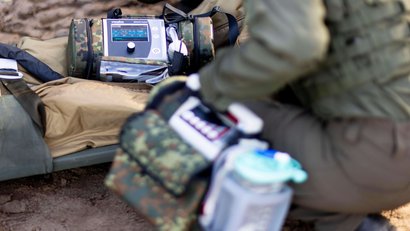 Ein Soldat kniet neben einer Trage im Freien. Auf der Trage liegt ein mobiles Beatmungsgerät in Tarnoptik.
