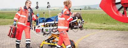 Notfallsanitäterinnen bringen einen Patienten mit Beatmungsgerät auf einer Fahrtrage zum Rettungshubschrauber.