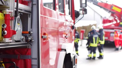 Im Vordergrund ist ein geöffnetes Feuerwehrfahrzeug. Im Hintergrund stehen mehrere Einsatzkräfte in Schutzkleidung.