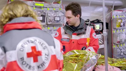 Zwei Einsatzkräfte versorgen in einem mobilen Behandlungszelt eine Person mit einer Rettungsdecke. Im Hintergrund sind medizinische Geräte und Notfallausrüstung.