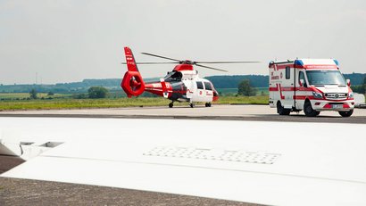 Ein Rettungshubschrauber und ein Rettungswagen stehen auf einem Flugfeld. Die Szene zeigt eine Notfallversorgung aus der Luft und am Boden.
