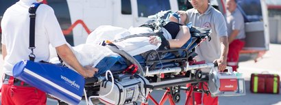 Zwei Rettungskräfte bringen eine verletzte Person auf einer Trage zum Krankenwagen. Die Person ist an medizinische Geräte angeschlossen und bekommt Sauerstoff.