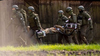 Soldaten in Uniform tragen eine verletzte Person auf einer Trage durch unwegsames Gelände. Ein Beatmungsgerät ist mit dabei.